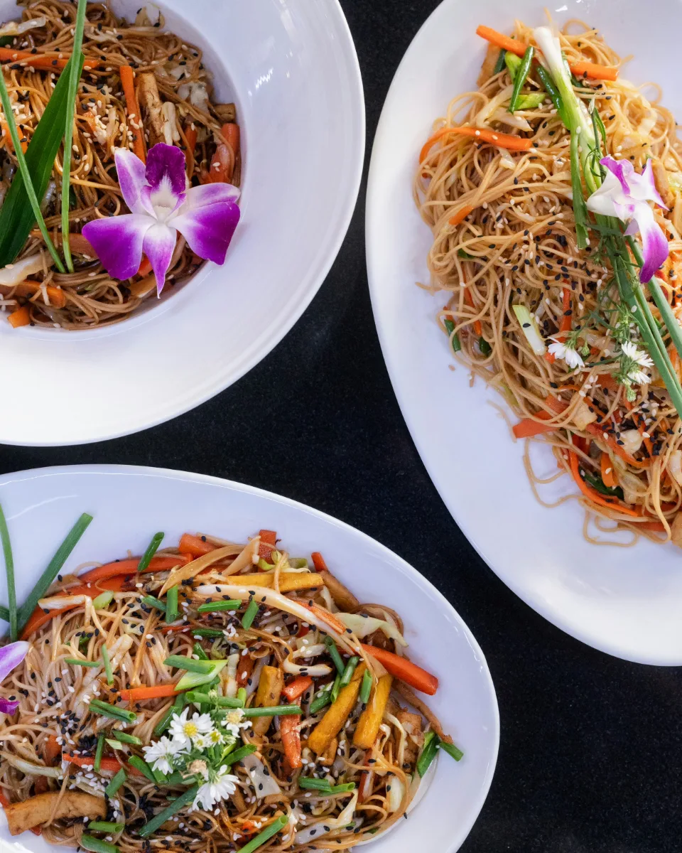 Three plates of stir-fried rice noodles with vegetables, tofu and orchids, seen from above